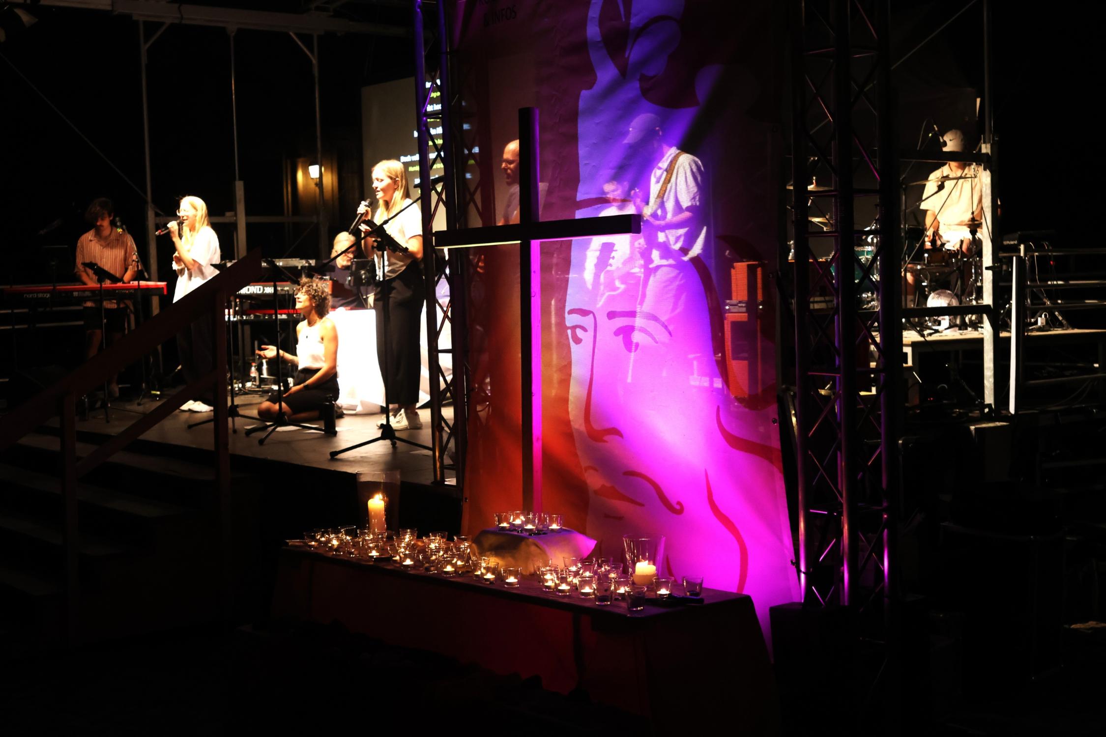 Illuminiertes Holzkreuz vor einem Banner des Heinrichsfests, davor Kerzen, im Hintergrund die Bühne