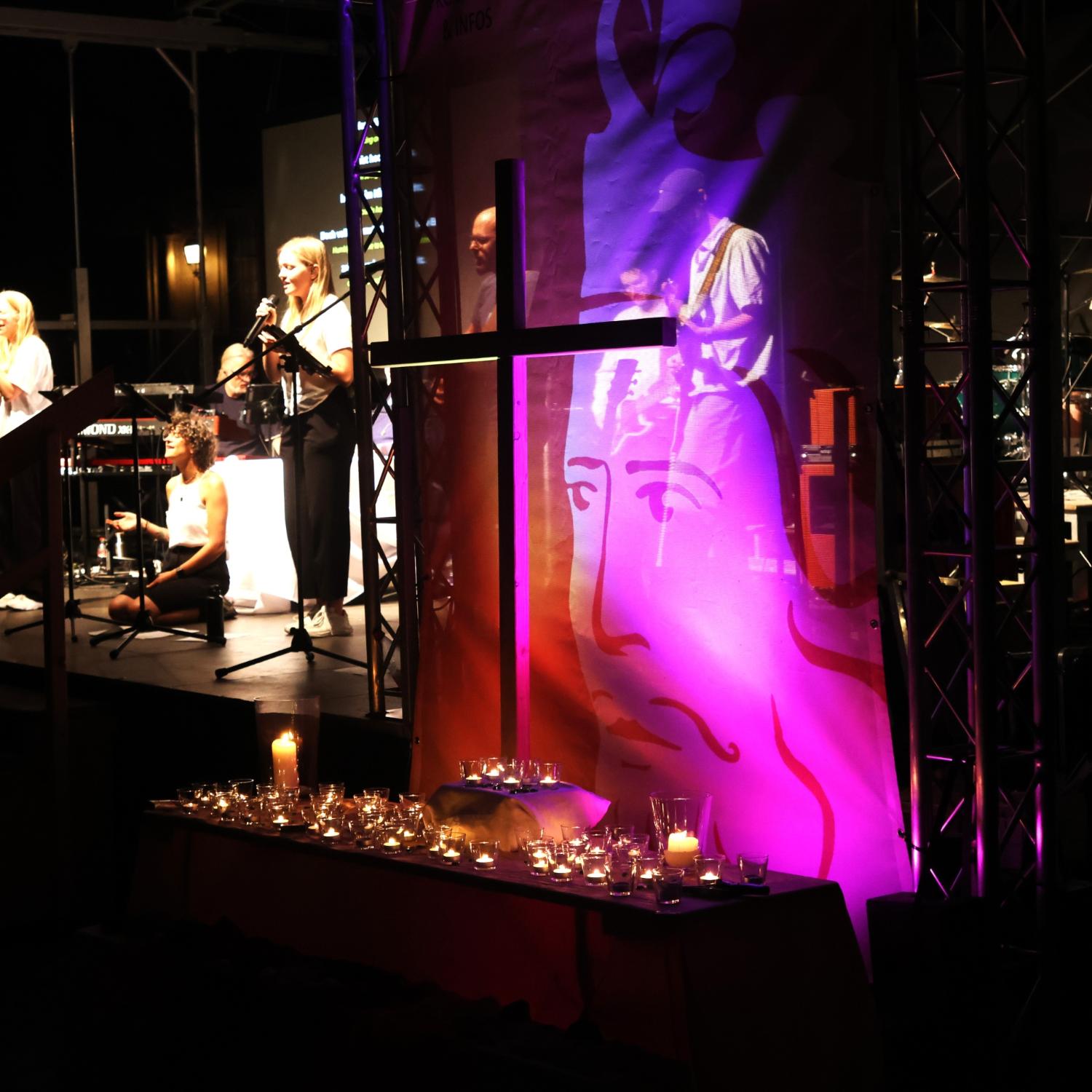 Illuminiertes Holzkreuz vor einem Banner des Heinrichsfests, davor Kerzen, im Hintergrund die Bühne