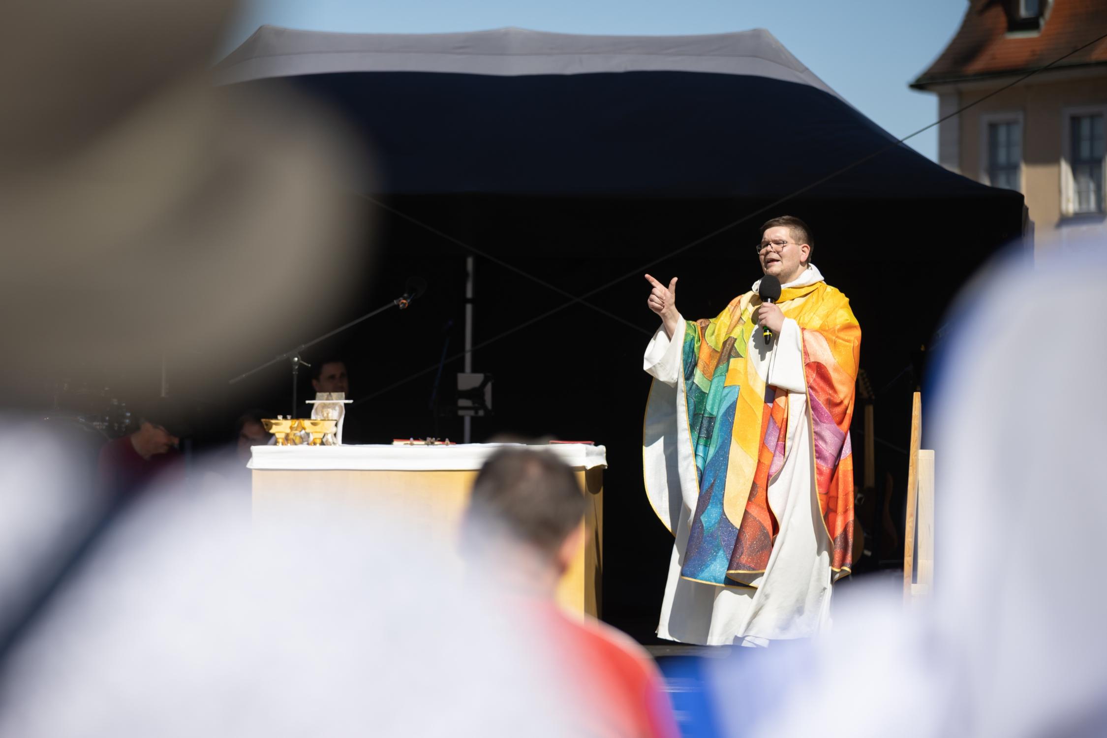 Pfarrer Grasser in buntem Messgewand mit Mikrofon und gestikulierend auf der Bühne, im Vordergrund sehr unscharf Gottesdienstteilnehmende