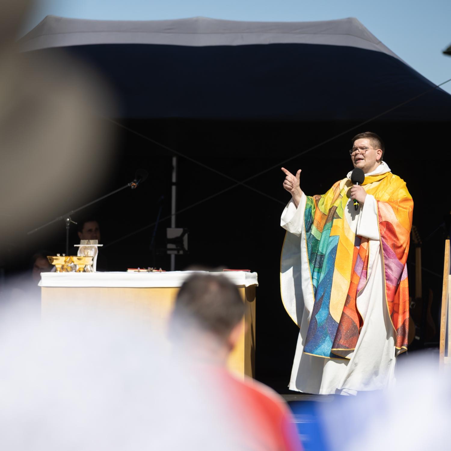 Pfarrer Grasser in buntem Messgewand mit Mikrofon und gestikulierend auf der Bühne, im Vordergrund sehr unscharf Gottesdienstteilnehmende