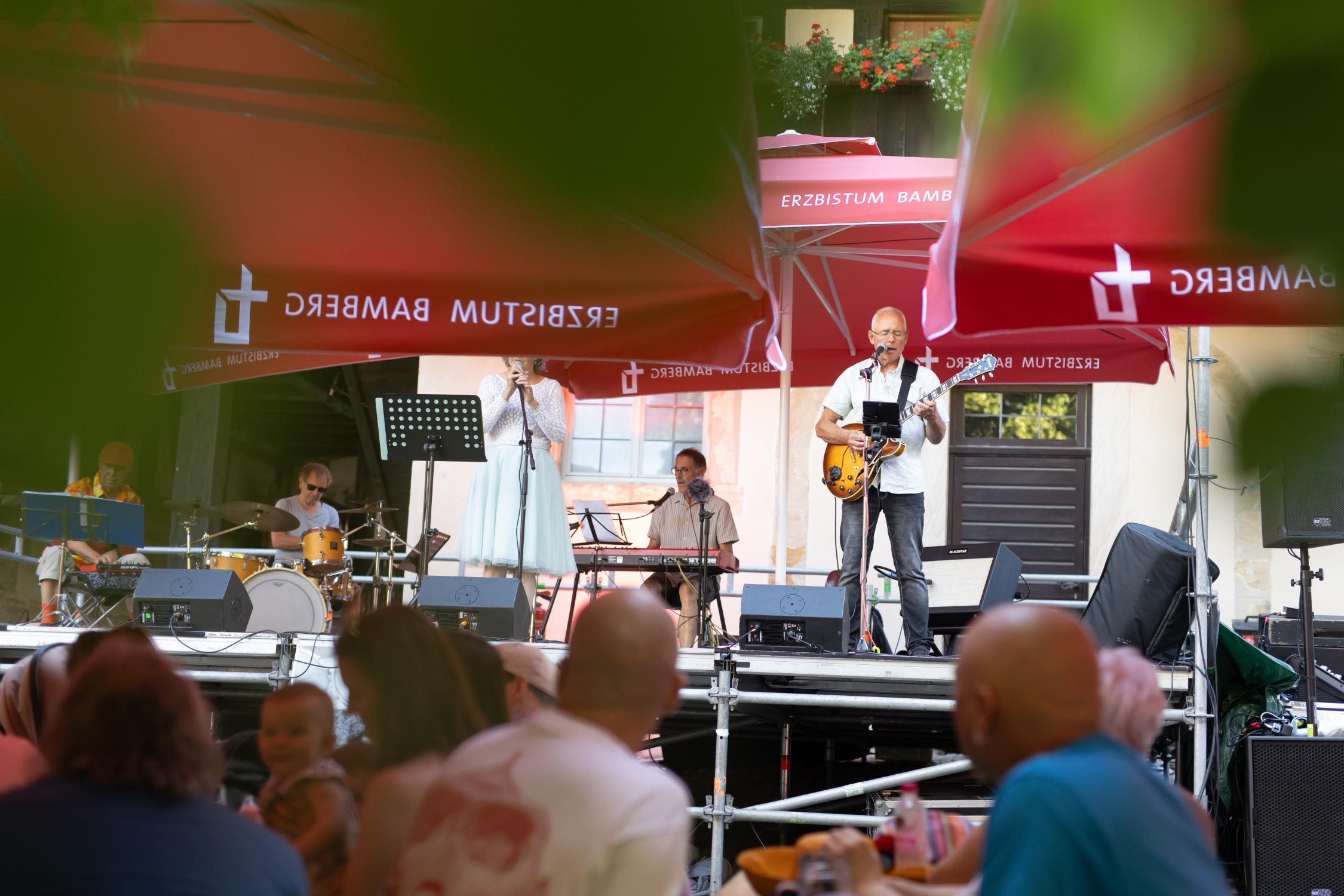 Bühne mit den Musikerinnen und Musikern, im Vordergrund unscharf Menschen an Biertischen